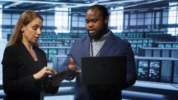 Teamworking colleagues doing checkup, inputting data on laptop of needed server hub fixes. Coworkers working together in data center housing advanced storage infrastructure, camera A photo