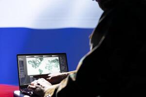 Russian ex KGB FSB operative engaged in surveillance mission using laptop, looking at world map. Intelligence officer isolated over Russia flag working for Kremlin regime monitoring zone of interest photo