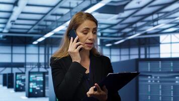 IT specialist looking over data center maintenance checklist, talking on phone in server farm. Employee surrounded by server racks talking during telephone call, camera A photo
