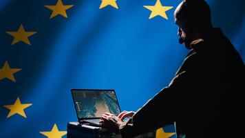 European Union military dispatcher looking at surveillance broadcast, relaying informations for troops using headset. Telecommunications operator over EU flag background, coordinating troops, camera B photo