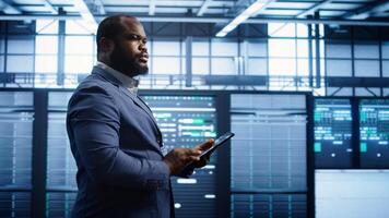 Technician walking in server room, doing checkup, inputting data on tablet of needed fixes. IT professional working in data center facility housing storage infrastructure, camera B photo