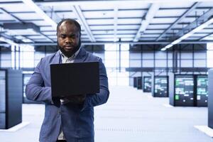 Technician walking in data center, monitoring system performance with laptop, identifying bottlenecks and implementing optimizations to improve efficiency. Server hub employee troubleshooting errors photo