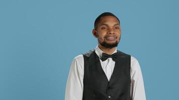 Friendly bellman porter with suit and tie waving at hotel guests, greeting people and offering excellent concierge services. Young employee saying hello to clients against blue background. Camera A. photo