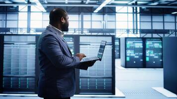African american man walking in high tech facility, using laptop to patch network issues leading to slow data transfer rates. Data center worker fixing errors affecting supercomputers, camera B photo
