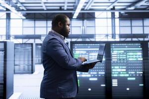 Employee in server hub using notebook device to monitor energy consumption across parts. IT professional in data center making sure infrastructure rigs are functioning optimally photo