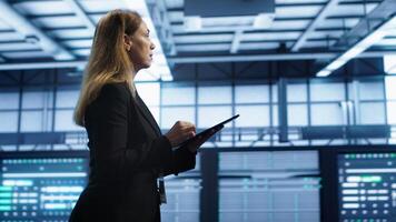 Engineer in server hub checking recovery plan on tablet, monitoring rackmounts energy consumption. Woman in data center making sure rigs are functioning optimally, doing routine disk checks, camera A photo