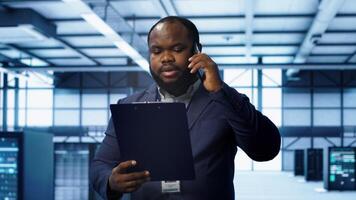 IT professional looking over data center checkup checklist paperwork, talking on phone in server farm. African american man surrounded by server rigs, chatting during telephone call, camera A photo