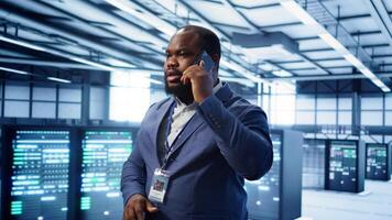 IT support worker in data center talking in phone call, doing yearly checkup. On field engineer chatting with manager in server hub during telephone call, monitoring servers infrastructure, camera B photo