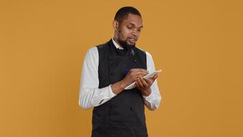 Skilled waiter with apron takes meal orders from clients on a notebook, writing down important information for customer satisfaction. Caterer butler working in service oriented industry. Camera B. photo