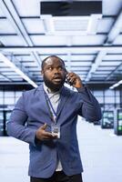 IT supervisor overseeing operations in cutting edge data center, coordinating tasks during telephone call. African american man in server room managing systems and communicating via smartphone photo