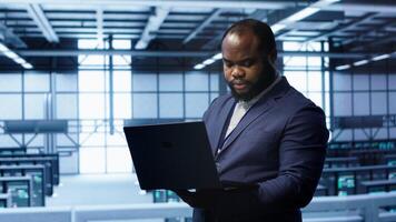 African american IT operator doing maintenance using laptop to prolong data center equipment life span. IT professional in server hub workspace monitoring server mainframes using notebook, camera A photo