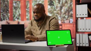 Businessman examines annual sales reports on documents next to a green screen tablet display, doing an accounting analytics plan. Entrepreneur working remotely from home office. Camera B. photo