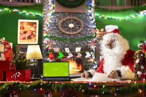 Santa Claus sitting next to copy space screen on device and reading letters from children around the world. Old man Saint Nicholas spreading his festive cheer for the holiday period. photo