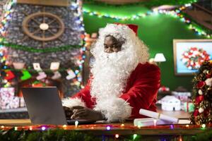Father Christmas is navigating the web checking on children wish lists using his laptop. Festive North Pole cabin is filled with holiday cheer and online activity this x mas, checking the world map. photo