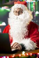 Father Christmas is navigating the web checking on children wish lists using his laptop. Festive North Pole cabin is filled with holiday cheer and online activity this x mas, checking the world map. photo