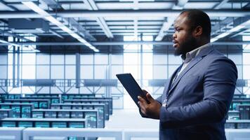 IT operator doing maintenance using tablet to prolong data center equipment life span. IT professional in server hub workspace monitoring server mainframes using digital device, camera B photo