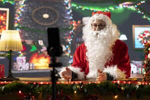 Father Christmas records a vlog talking to his online audience about the magic of Christmas. Santa Claus in a cozy festive cabin connects with subscribers through social media during wintertime. photo