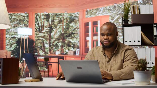 Entrepreneur sitting at his desk working on laptop with wireless technology for remote job efficiency. Analyzing data and developing strategies, successful telecommuting from home office. Camera B. photo