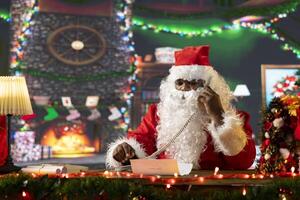 Santa Claus reading letters and calling helpers on landline phone call, in his festive decorated North Pole. Spreading holiday cheer with Christmas lights and decorations, magic of gift giving. photo