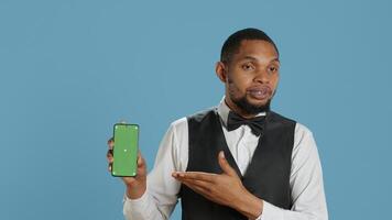 Hotel worker bellhop points at phone with isolated mockup display, showing green screen against blue background in studio. Hotelier doorman greeting resort guests at reception. Camera A. photo