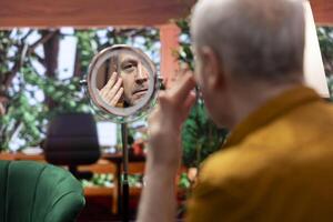 Elderly man looking in the mirror at his wrinkles and aging skin while he does his daily routine for grooming and skincare. Applying rejuvenating cream and lotion for hydration. photo