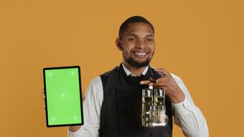 Male restaurant staff showing tablet with green screen against yellow background, carrying the cruet set with salt and pepper shakers used in serving clients. Service oriented job. Camera A. photo