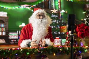 Saint Nicholas dressed in his red suit filming a diary in his decorated North Pole cabin. White bearded influencer shares holiday traditions with his online audience in a festive broadcast. photo