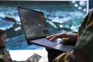 Intelligence specialist monitoring a world map and surveillance imagery on laptop, using the satellite radar system with cctv. Soldiers manage reconnaissance activity in army command post. photo