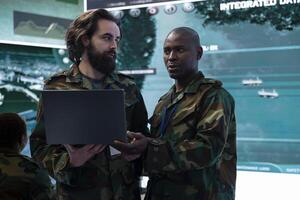 Diverse team of IT operators comparing real time battlefield data from a laptop to a big screen, gather intelligence for future field missions. Collecting information from CCTV satellite surveillance. photo