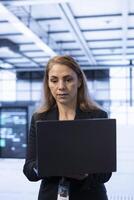 Programmer in server hub workplace installing newly ordered storage unit, checking parameters with laptop. IT specialist in data center holding notebook, replacing storage mainframes components photo