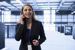 Smiling person overseeing operations in cutting edge data center, coordinating tasks during telephone call. Happy IT specialist in server hub managing systems and communicating via smartphone photo