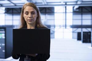 Data center programmer ensuring safety and security of database using laptop. Woman in server farm preventing unauthorized access and suspicious activities posing risks for business photo