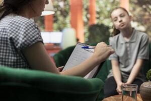Psychologist taking notes of teenager behavioral progress during therapy session, helping her with encouragement to overcome personal troubles and hormonal shifts. Therapist writing complaints. photo