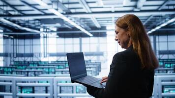Computer scientist doing yearly maintenance using laptop to prolong data center equipment life span. IT specialist in server hub facility monitoring server infrastructure using device, camera B photo