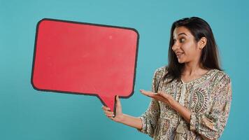 Smiling indian woman holding red speech bubble sign of empty copy space for message. Joyous person presenting thought bubble cardboard used as promotion concept, studio background, camera A photo