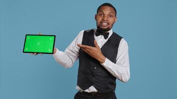 Doorkeeper bellboy poses with green screen on tablet at the hotel reception, working in hospitality industry and greeting customers. Lodging resort personnel shows chroma key. Camera B. photo