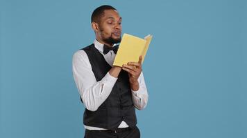 Hotel concierge in tuxedo reading a novel story book in studio, enjoying literature and lecture hobby. Hospitality industry staff working in five star lodging resort, bellhop receptionist. Camera B. photo