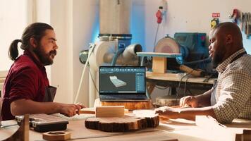 Artisan and apprentice comparing wood art piece with schematic displayed on CAD software on laptop. Woodworking experts in studio crosschecking wooden object with notebook blueprint, camera A photo