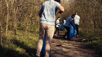 Team of volunteers cleaning the forest area from garbage and plastic, collecting rubbish with tongs and bags. Activists doing voluntary work to preserve natural habitat, illegal dumping. Camera B. photo