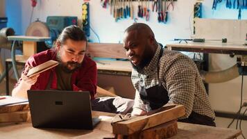 Carpenter and colleague verifying finished wood art product, comparing with blueprint. Artisan and apprentice inspecting wooden object for damages based on schematics on laptop screen, camera A photo