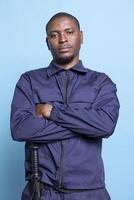 Portrait of african american guard posing with arms crossed against blue background, wearing his professional uniform with confidence and pride. Serious armed agent works on patrol service. photo