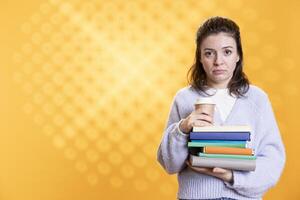 Portrait of woman with worried look on face carrying stack of books and coffee cup, studio background. Student feels anxious before university assessment, holding textbooks, drinking caffeine beverage photo