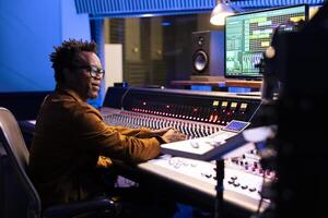 African american sound designer operating audio software to edit tracks and control panel board to mix and master the tunes. Professional music producer creating new songs, motorized faders. photo