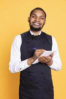 Professional caterer writing clients orders on notepad for the bill, takes notes of desired food against yellow background. Waiter with apron, notebook service for customer satisfaction. photo