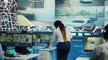 African american manager provides information for delivery parcels to her diverse team, people monitoring traffic on routes. Order tracking in control room, satellite maps with coordinates. Camera B. photo