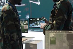 Team of diverse troopers examining satellite CCTV footage on a tablet, collecting information for a reconnaissance mission in command center. Soldiers engineers operating high tech gear. photo