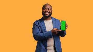 Smiling influencer presenting green screen mobile phone, isolated over studio background. Joyful BIPOC person holding copy space chroma key smartphone used for advertising brands, camera B photo