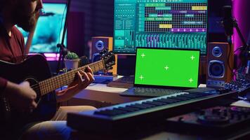Musician beginner learning accords to play acoustic guitar by watching tutorials and online lessons, laptop shows greenscreen chromakey. Artist practicing guitar skills in home studio. Camera A. photo