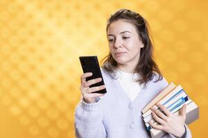 Smiling woman holding pile of books, texting friends with smartphone, conveying joy of reading concept, studio background. Cheerful person with novels in arms talking with mates on mobile phone photo