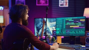 Music producer operating with electronic setup of home studio, recording acoustic guitar sounds. Artist navigating signal processing activity, utilizing digital audio workstation at desk. Camera A. photo
